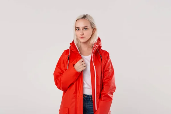 Glimlachend blond meisje in een rode regenjas en wit t-shirt staande geïsoleerd over witte achtergrond. Maak je klaar voor slecht weer. — Stockfoto