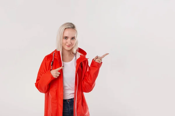 Chica rubia bastante sonriente con un impermeable rojo con capucha apuntando con un dedo al espacio vacío en el lado derecho de pie aislado sobre fondo blanco. Lugar para la publicidad —  Fotos de Stock