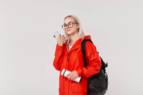 Blond flicka i en röd regnrock och glasögon reser med svart ryggsäck och vit termisk Mugg i kall säsong. Regnperioden. — Stockfoto