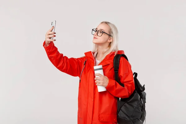 Menina loira em uma capa de chuva vermelha e óculos fazendo selfie com um smartphone viajando com mochila preta e caneca térmica branca na estação fria. Temporada chuvosa . — Fotografia de Stock