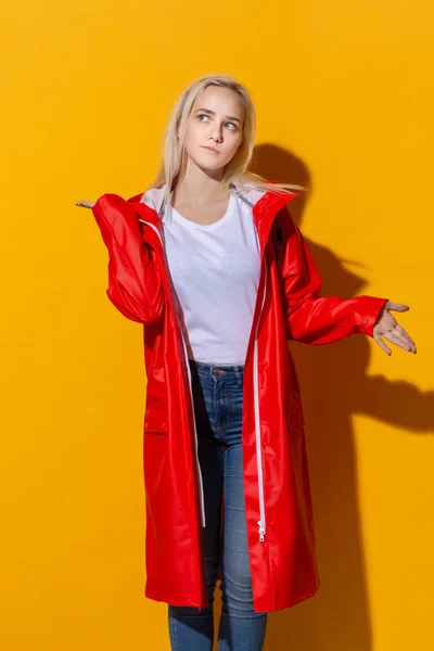 Linda chica rubia con un impermeable rojo y una camiseta blanca de pie aislada sobre fondo amarillo. Ser brillante en el mal tiempo . — Foto de Stock