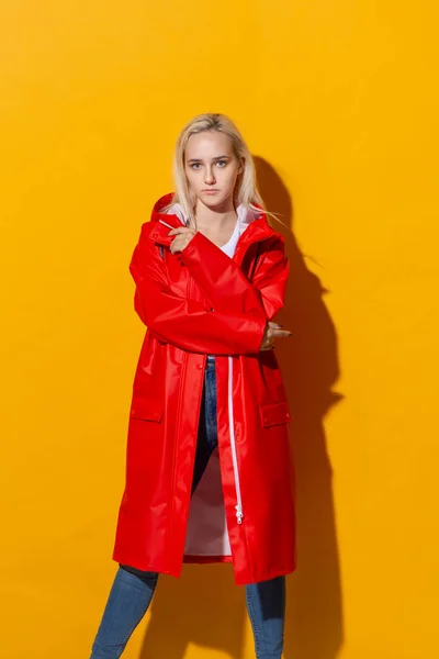 Pretty blond girl in a red raincoat and white t-shirt standing isolated over yellow background. Be bright in bad weather. — Stock Photo, Image