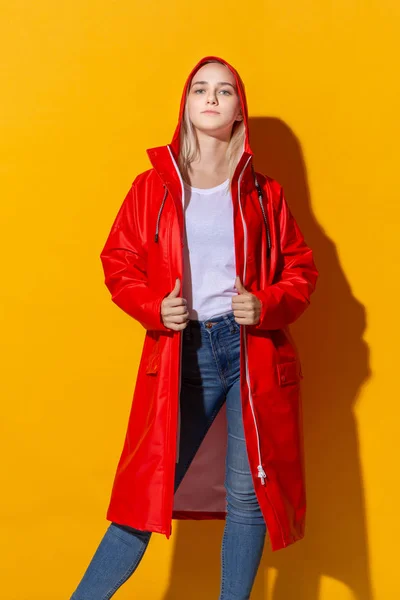 Pretty blond girl in a red raincoat and white t-shirt standing isolated over yellow background. Be bright in bad weather. — Stock Photo, Image