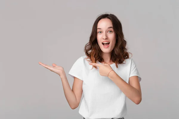 Hübsche brünette Frau in einem weißen T-Shirt, das eine Handfläche mit leerem Raum für Produktplatzierung hält und isoliert vor grauem Hintergrund steht. Kopierraum der Werbung. — Stockfoto