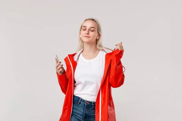 Jolie fille blonde portant un imperméable rouge et un t-shirt blanc dansant en écoutant de la musique avec des écouteurs sans fil. Concept de profiter de la vie . — Photo