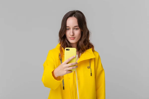 Mulher morena bonita em uma capa de chuva amarela com um smartphone de verificação de capuz para notificações isoladas sobre fundo cinza. Indo para um passeio em um dia frio chuvoso — Fotografia de Stock