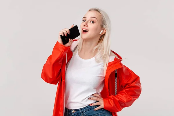 Menina muito loira vestindo capa de chuva vermelha e branco t-shirt dançando ouvir música com fones de ouvido sem fio e cantando músicas com smartphone. Conceito de desfrutar da vida . — Fotografia de Stock