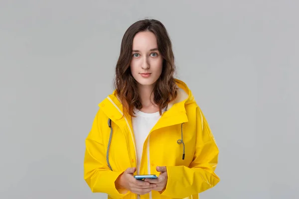 Mujer morena bonita en un impermeable amarillo con un teléfono inteligente de comprobación de capucha para las notificaciones aisladas sobre fondo gris. Voy a dar un paseo en un día frío lluvioso —  Fotos de Stock