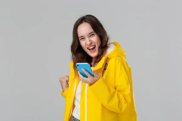 Mujer morena bonita en un impermeable amarillo expresando emoción de éxito sosteniendo teléfono inteligente aislado sobre fondo gris. Concepto de éxito . — Foto de Stock