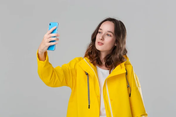 Mujer morena bonita en un impermeable amarillo haciendo selfie con teléfono inteligente aislado sobre fondo gris. Voy a dar un paseo en un día frío lluvioso — Foto de Stock