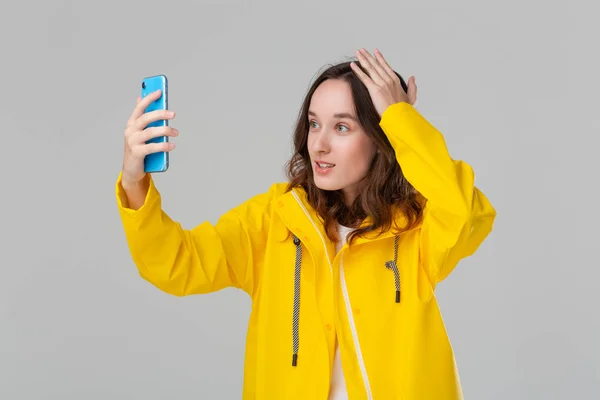 Sarı yağmurluklu güzel esmer kadın gri arka planda izole edilmiş akıllı telefondan sohbet ediyor. İletişim kavramı. — Stok fotoğraf