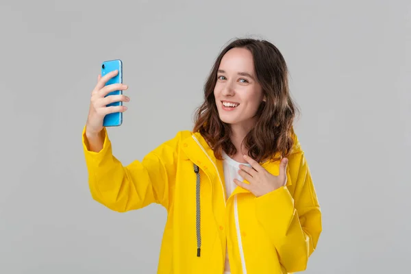 Mulher morena bonita em uma capa de chuva amarela fazendo selfie com smartphone isolado sobre fundo cinza. Vou dar uma volta num dia frio e chuvoso. Faça dias nebulosos mais brilhantes — Fotografia de Stock