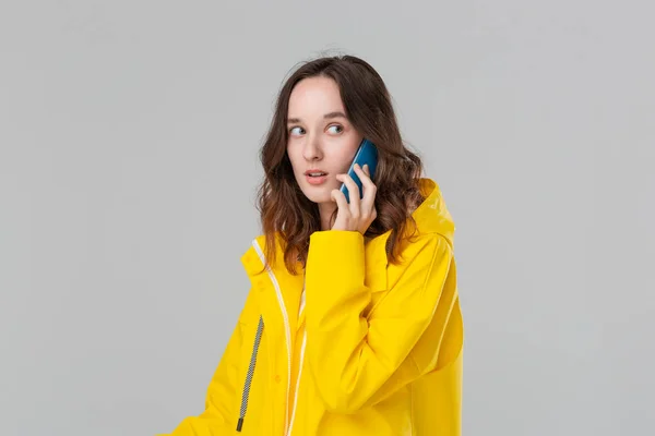 Pretty brunette woman in a yellow raincoat looking worried talking on the mobile phone isolated over grey background. Concept of communication. — ストック写真