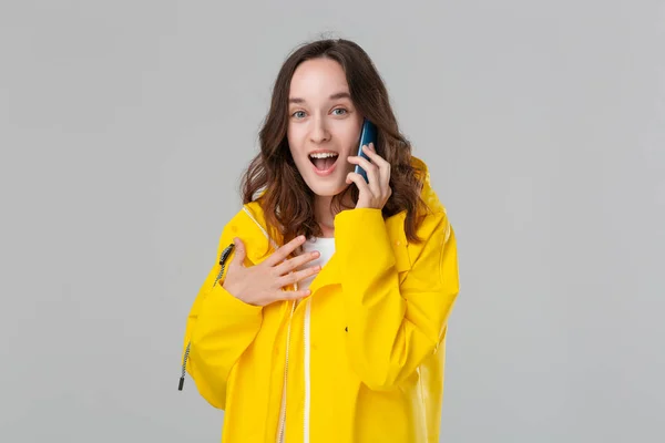 Mujer morena bonita en un impermeable amarillo que se ve feliz hablando en el teléfono móvil aislado sobre fondo gris. Concepto de comunicación . —  Fotos de Stock