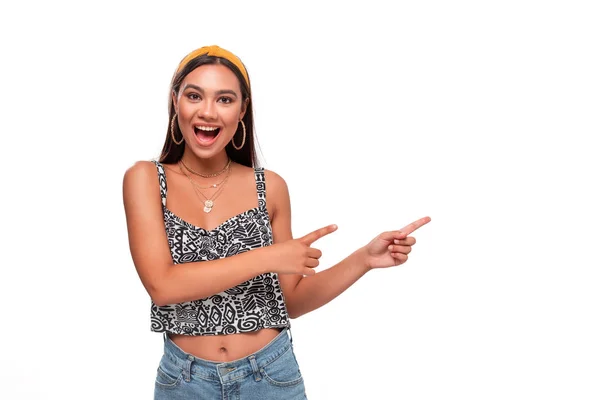 Pretty african-american teenager in a headband, sleeveless t-shirt and jeans pointing at the empty space in a right side of a frame standing isolated over white background. Copy space of advertising. — Stock Photo, Image