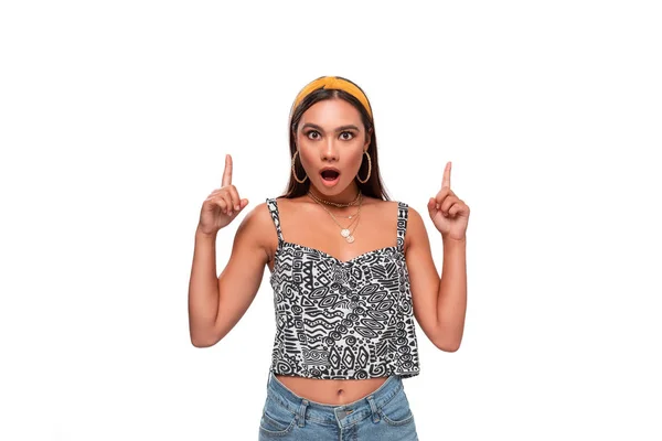 Mooie Afrikaans-Amerikaanse tiener in een hoofdband, mouwloos t-shirt en jeans wijzend naar de lege ruimte in een bovenzijde van een frame dat geïsoleerd staat over een witte achtergrond. Kopieerruimte van reclame. — Stockfoto