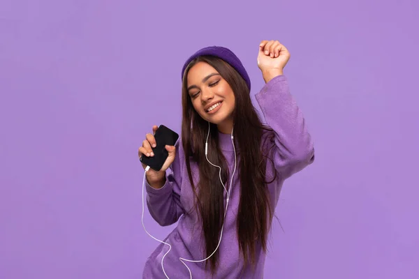 Şapkalı ve kazaklı güzel bir Afro-Amerikalı kız. Cep telefonuyla müzik dinliyor. Kulaklıkları kablosuz ve mor arka planda dans ediyor. Hayatın tadını çıkarıyorum..