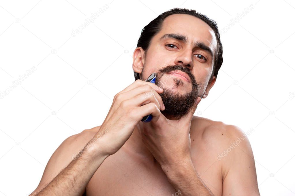 Handsome man with mustache uses electric shaver to trim his beard standning bare isolated over white background. Concept of morning treatment and shaving. Time to trim your beard. Morning routine