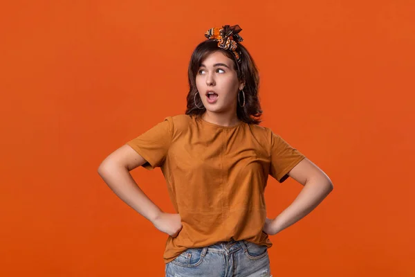 Pretty brunette woman in a t-shirt and beautiful headband expressing emotion of surprise standing isolated over orange background.
