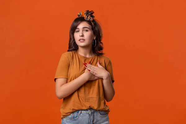 Pretty brunette woman in a t-shirt and beautiful headband expressing sincere feeling putting hand to heart standing isolated over orange background. Concept of sincerity