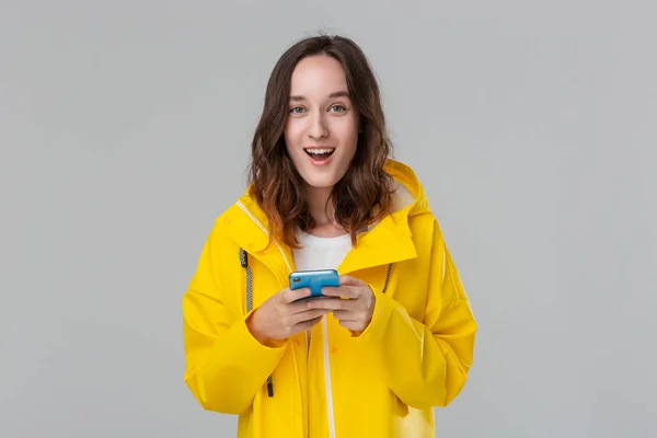 Mujer morena bonita en un impermeable amarillo se emociona leyendo notificación en el teléfono inteligente aislado sobre fondo gris. Concepto de comunicación . — Foto de Stock