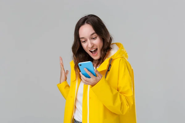 Pretty brunette woman in a yellow raincoat expressing emotion of success holding smartphone isolated over grey background. Concept of success. — Stock Photo, Image