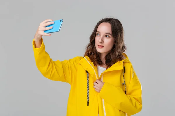 Mujer morena bonita en un impermeable amarillo haciendo selfie con teléfono inteligente aislado sobre fondo gris. Voy a dar un paseo en un día lluvioso y frío. Hacer los días brumosos más brillantes — Foto de Stock