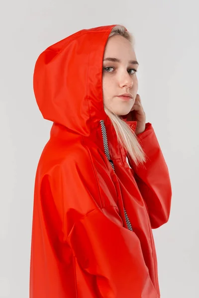 Mooi blond meisje krijgt op de motorkap van een rode regenjas geïsoleerd over een witte achtergrond. Het begint te regenen.. — Stockfoto