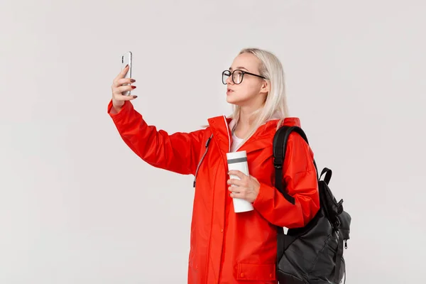 Menina loira em uma capa de chuva vermelha e óculos fazendo selfie com um smartphone viajando com mochila preta e caneca térmica branca na estação fria. Temporada chuvosa . — Fotografia de Stock