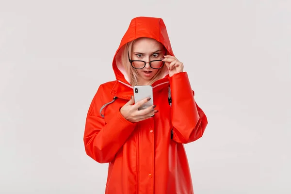 Mujer rubia en un impermeable rojo con capucha y gafas revisando smartphone para las notificaciones que expresan emoción de sorpresa aislada sobre fondo blanco. Voy a dar un paseo en un día frío lluvioso —  Fotos de Stock
