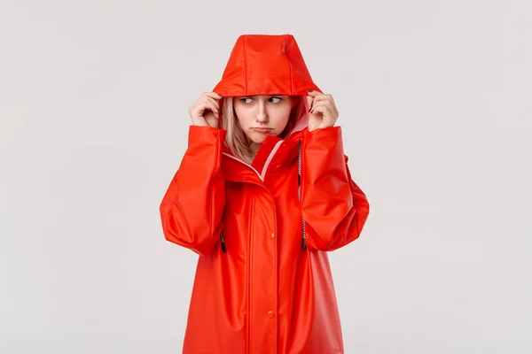 Blond girl in a red raincoat with a hood is freezing standing isolated over white background. Are you ready for cold bad weather?