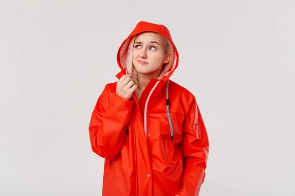 赤いレインコートを着たブロンドの女の子は、白い背景の上に孤立した雨が立ち始めたかどうかを確認するフードを持っています。寒い悪天候の準備はできていますか? — ストック写真