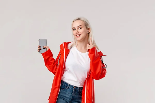 Une fille blonde portant un imperméable rouge et un t-shirt blanc dansant en écoutant de la musique avec des écouteurs sans fil. Concept de profiter de la vie . — Photo