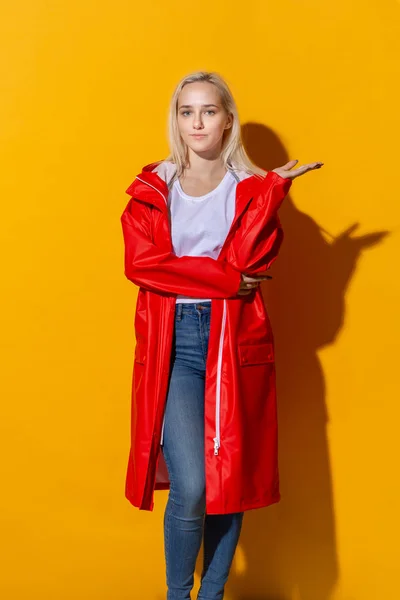 Chica rubia con un impermeable rojo y una camiseta blanca de pie aislada sobre fondo amarillo. Ser brillante en el mal tiempo . — Foto de Stock