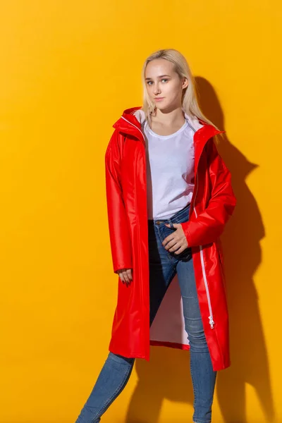 Blond girl in a red raincoat and white t-shirt standing isolated over yellow background. Be bright in bad weather. — Stock Photo, Image