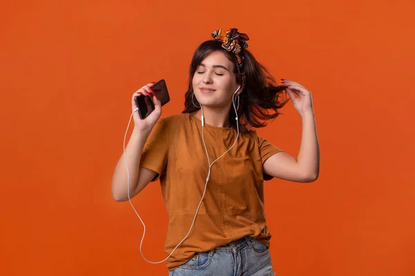 Bir t-shirt ve güzel kafa bandı turuncu arka plan üzerinde izole kablolu kulaklıklar ile müzik dinlerken dans Güzel esmer kadın. Hayatın tadını çıkarmak