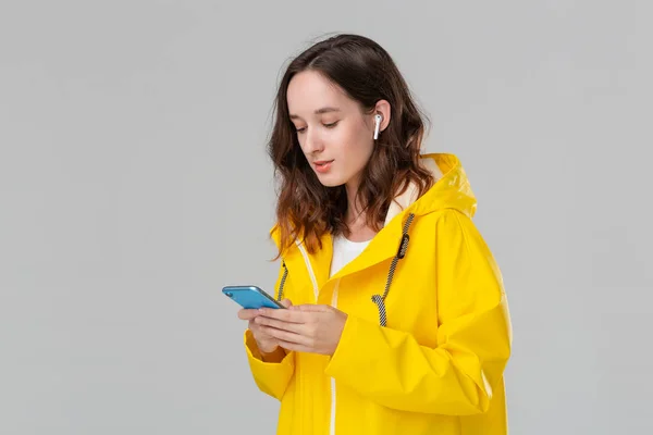 Mujer morena bonita en un impermeable amarillo está leyendo notificación en el teléfono inteligente aislado sobre fondo gris. El modelo usa auriculares inalámbricos. Concepto de comunicación . — Foto de Stock