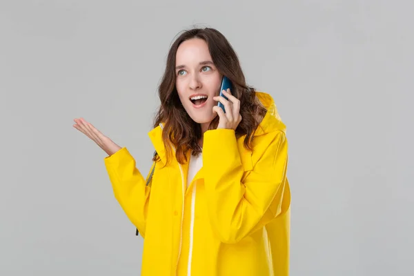 灰色の背景に隔離されたスマートフォンで幸せな話を見て黄色のレインコートの美しいブルネットの女性。コミュニケーションの概念. — ストック写真