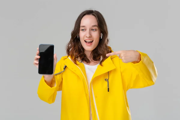 Mujer morena bonita en un impermeable amarillo que muestra la pantalla de un teléfono móvil aislado sobre fondo gris. El modelo usa auriculares inalámbricos. Lugar para la publicidad — Foto de Stock