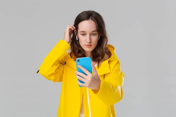 Mulher morena bonita em uma capa de chuva amarela está fazendo selfie com o smartphone isolado sobre fundo cinza. Modelo usa fones de ouvido sem fio. Fotografia móvel . — Fotografia de Stock
