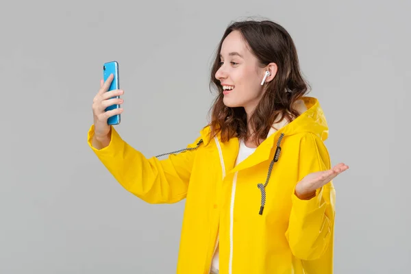 Pretty brunette woman in a yellow raincoat video chatting on the smartphone isolated over grey background. Concept of communication.