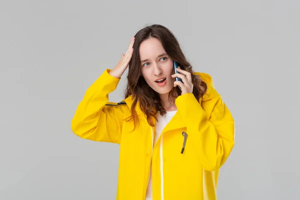 Sarı yağmurluk giymiş güzel esmer kadın gri arka planda izole edilmiş akıllı telefondan şaşkın şaşkın konuşuyor. İletişim kavramı. — Stok fotoğraf
