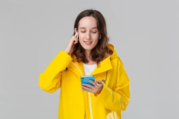 Mujer morena sonriente en un impermeable amarillo está leyendo notificación en el teléfono inteligente. El modelo usa auriculares inalámbricos. Concepto de comunicación . — Foto de Stock