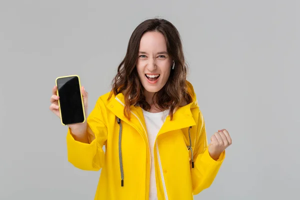 Mulher morena sorridente em uma capa de chuva amarela expressando emoção de sucesso mostrando tela do smartphone isolada sobre fundo cinza. Conceito de sucesso . — Fotografia de Stock