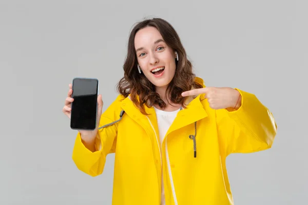 Mujer morena excitada en un impermeable amarillo que muestra la pantalla de un teléfono móvil aislado sobre fondo gris. El modelo usa auriculares inalámbricos. Lugar para la publicidad — Foto de Stock