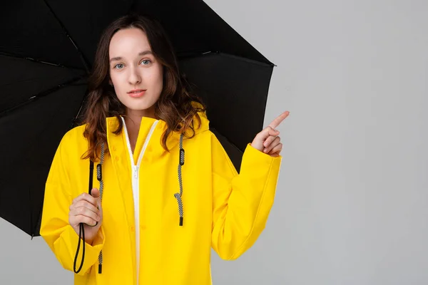 Linda chica morena escondida bajo paraguas negro vestida con impermeable amarillo y señalando con un dedo el espacio vacío para la publicidad . —  Fotos de Stock