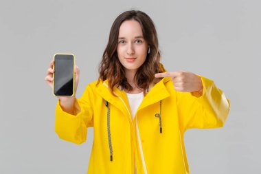 Sarı yağmurluklu esmer kadın gri arka planda izole bir cep telefonu ekranı gösteriyor. Model kablosuz kulaklık takıyor. Reklam için yer