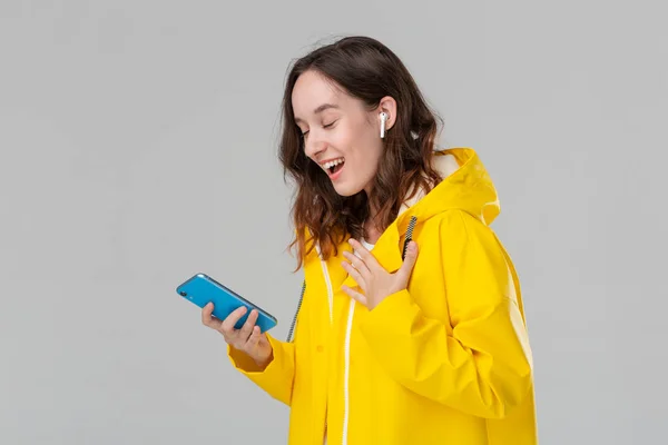Mujer morena sonriente en un impermeable amarillo está leyendo notificación en el teléfono inteligente. El modelo usa auriculares inalámbricos. Concepto de comunicación . — Foto de Stock