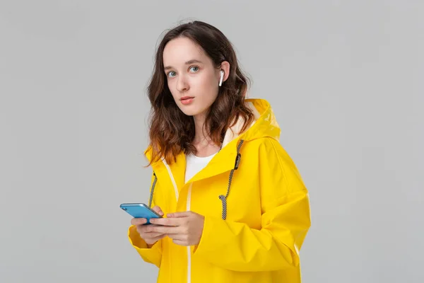 Mulher morena em uma capa de chuva amarela está lendo notificação no smartphone. Modelo usa fones de ouvido sem fio. Conceito de comunicação . — Fotografia de Stock