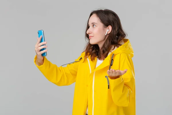 Mulher morena bonita em um vídeo capa de chuva amarela conversando no smartphone isolado sobre fundo cinza. Conceito de comunicação . — Fotografia de Stock